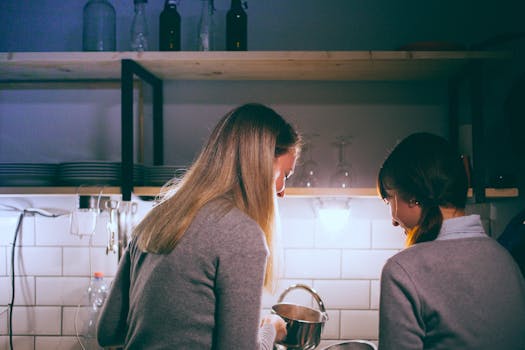 Back view of happy women with dishes standing near sink and having fun together in dark kitchen with light bulb